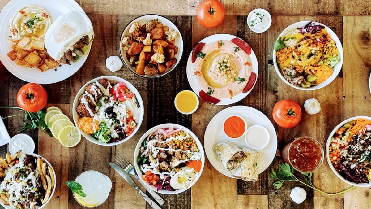 A wood table covered in AMBA bowls, wraps, fries, hummus and dips