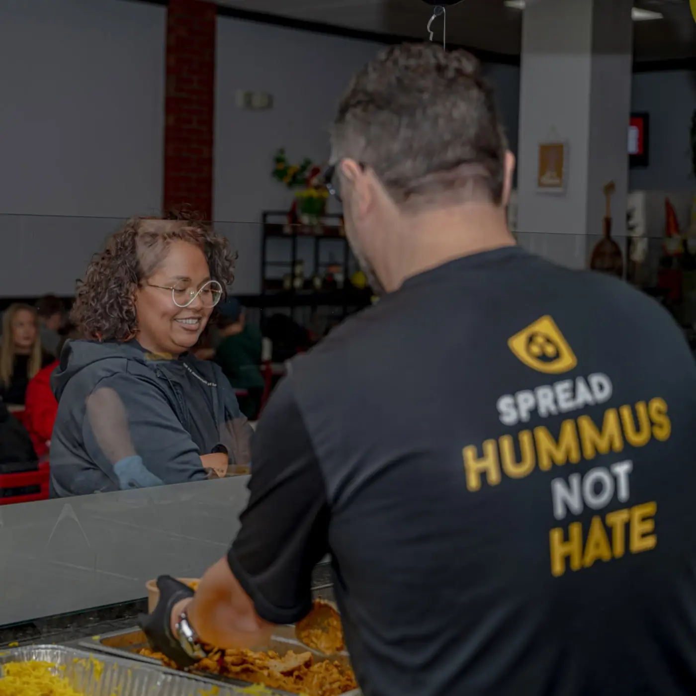 A crew member in a Spread Hummus Not Hate shirt building a bowl for a smiling guest at the counter